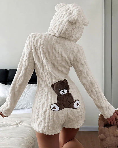A person shown from the back wearing a cream-colored, chunky knit hooded romper with a brown teddy bear applique on the back, standing in a bedroom setting.