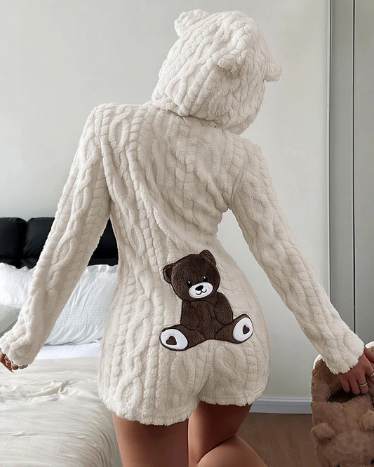 A person shown from the back wearing a cream-colored, chunky knit hooded romper with a brown teddy bear applique on the back, standing in a bedroom setting.