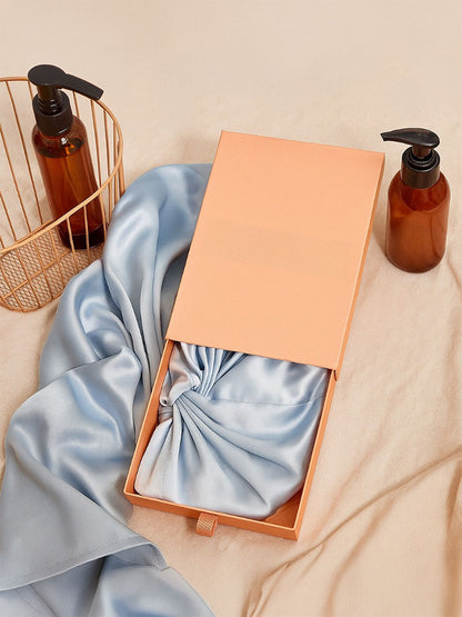 A peach-colored gift box with a satin ribbon tied around it, placed on a beige fabric surface next to two amber pump bottles and a gold wire basket.
