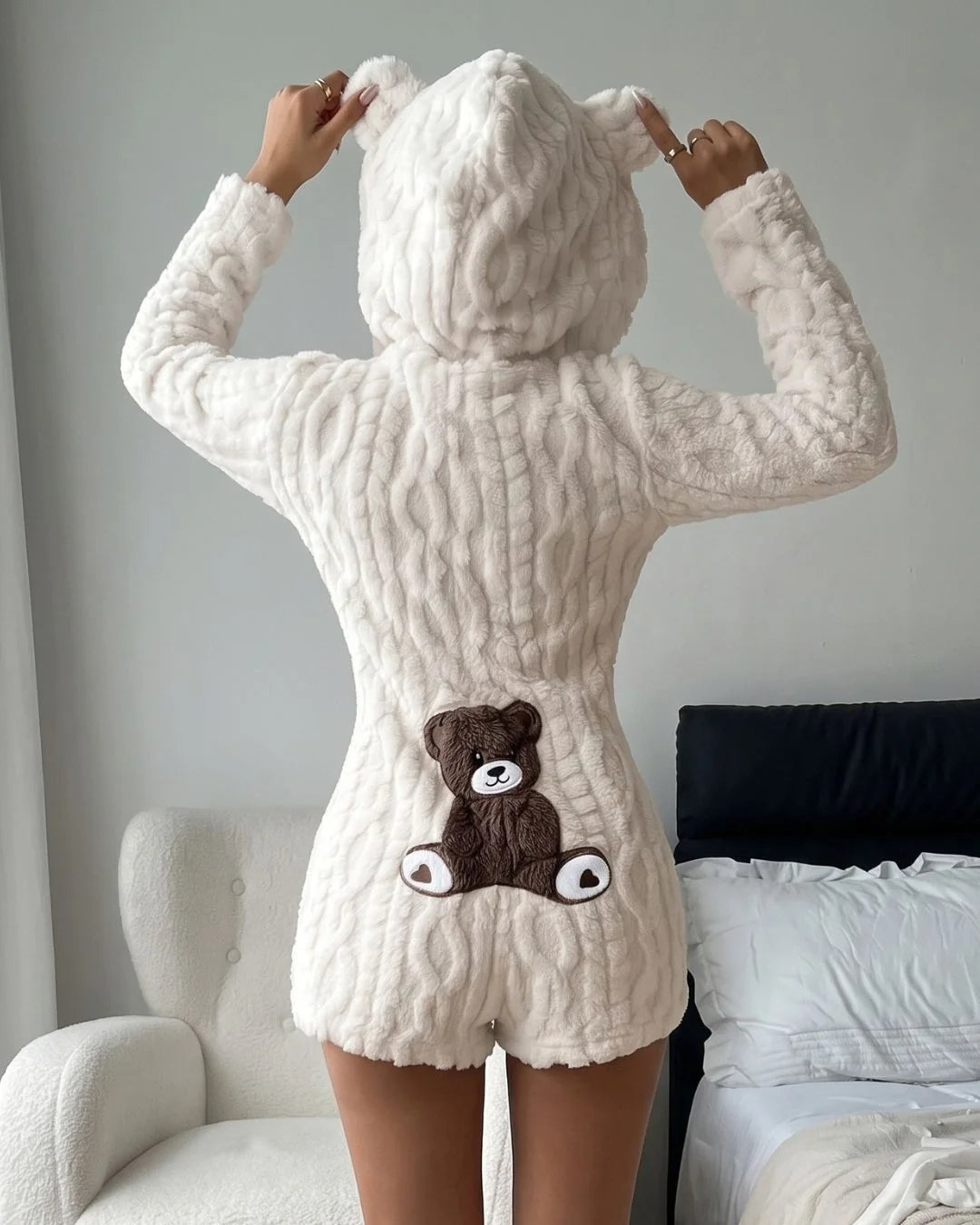 A person wearing a cream-colored, textured plush hooded romper with a brown teddy bear applique on the back, shown from behind in a bedroom setting.