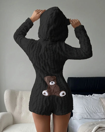 A person wearing a black textured hooded romper with a brown teddy bear applique on the back, standing in a bedroom setting.