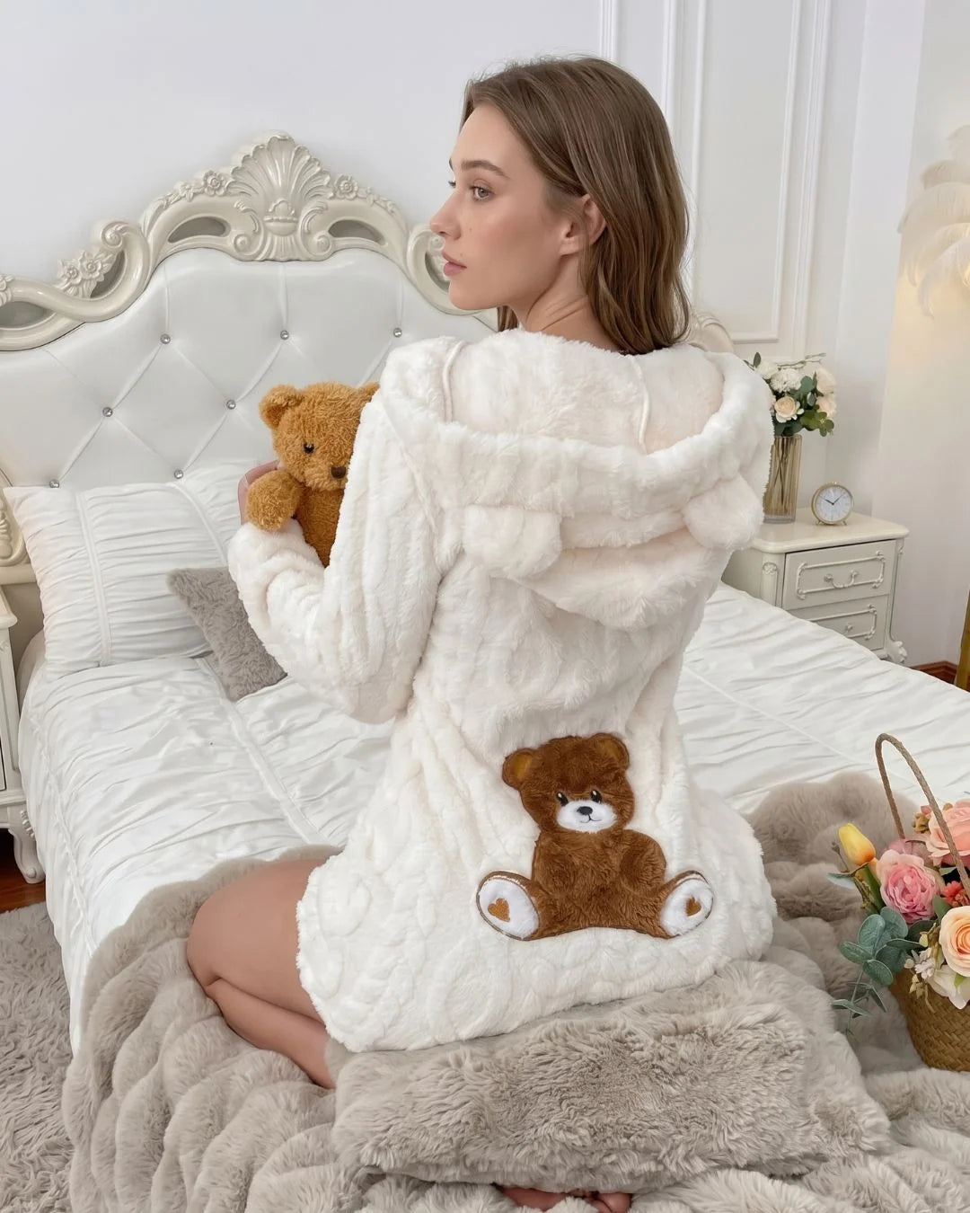 A woman sits on a bed wearing a fluffy cream hooded robe with a teddy bear applique on the front. She holds a small brown teddy bear, and the bedroom setting includes white bedding, a tufted headboard, and soft neutral decor.