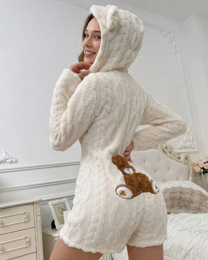 A woman wearing a cream-colored, cable-knit hooded romper with a brown teddy bear patch on the back, standing in a bedroom setting.