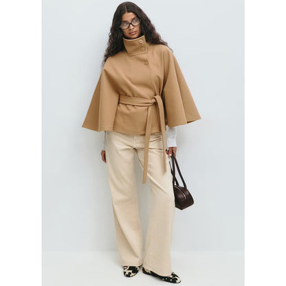 Woman wearing a beige cape over a white top and beige pants, holding a brown handbag against a light gray background.