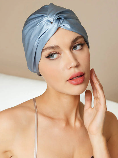 A woman wearing a light blue satin turban-style headwrap or wig cap, shown in a close-up portrait against a soft beige background.