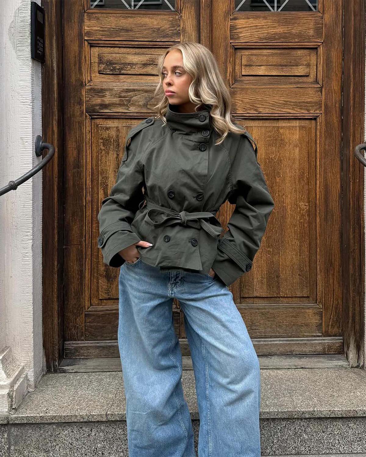 Woman wearing a green jacket and blue jeans standing in front of a wooden door.